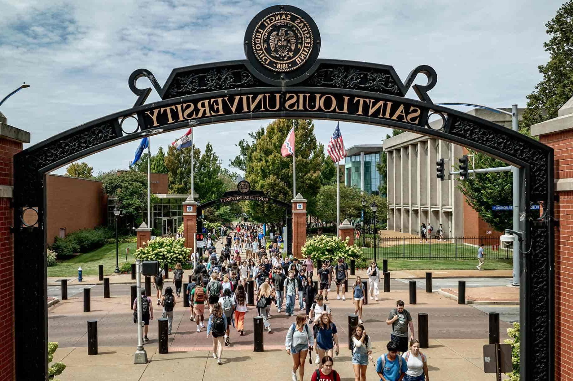 Students cross Grand Boulevard on a sunny afternoon. A pedestrian arch frames the view with “Saint Louis University” and the University seal visible at the top.